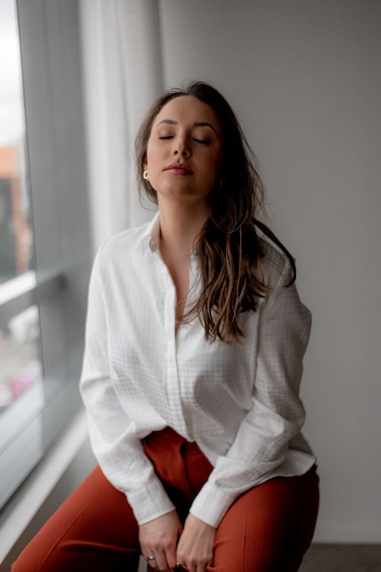 Mulher de olhos fechados em postura meditativa sentada junto à janela praticando mindfulness e yoga