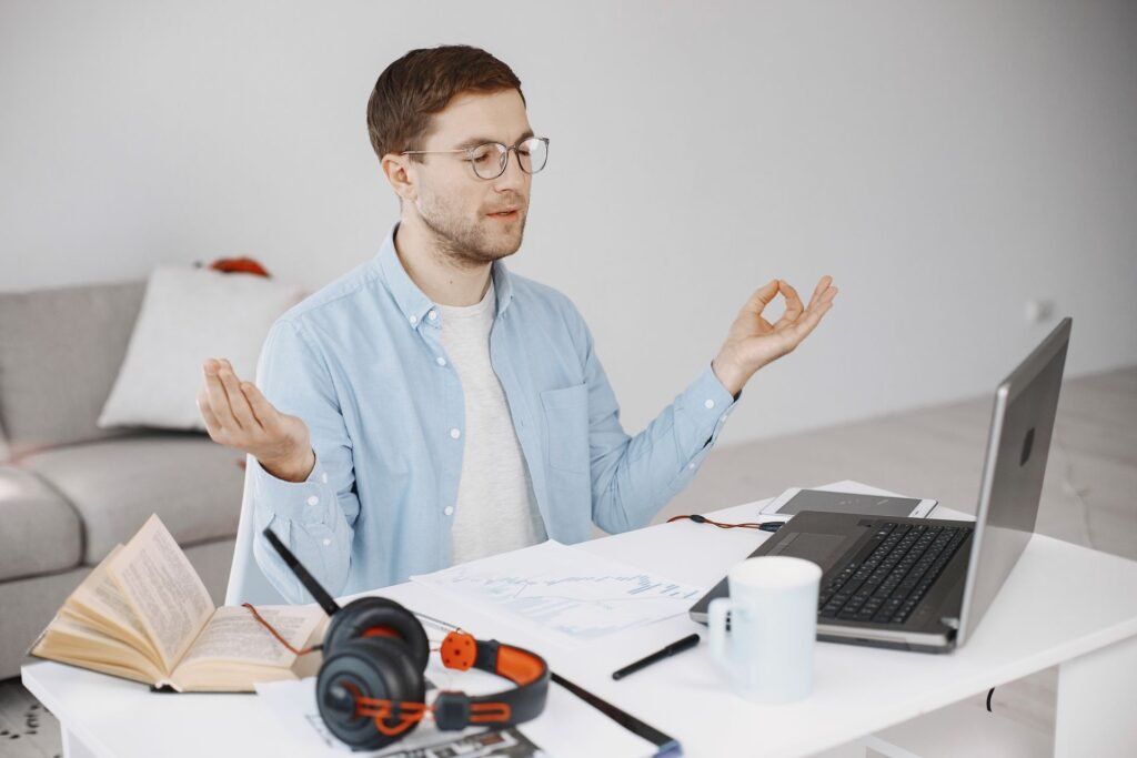 Homem de óculos meditando à mesa de trabalho com laptop e livros ao fundo