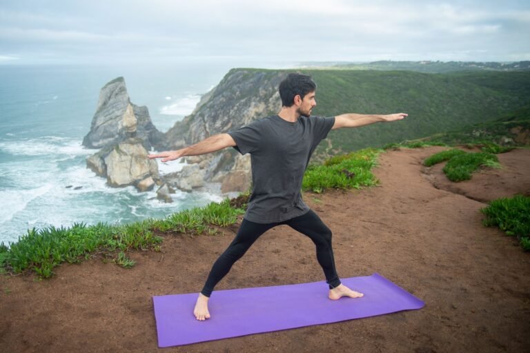 Homem praticando postura do Guerreiro II no tapete de yoga em falésia com vista para o oceano ilustrando yoga para homens como ferramenta de força mobilidade e performance atlética