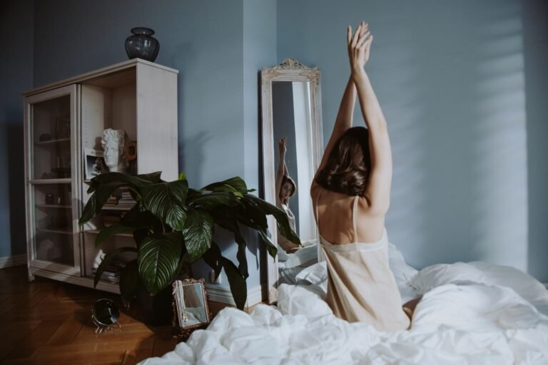 Mulher alongando os braços para cima, sentada na cama, em quarto claro e aconchegante durante a rotina de yoga para o sono