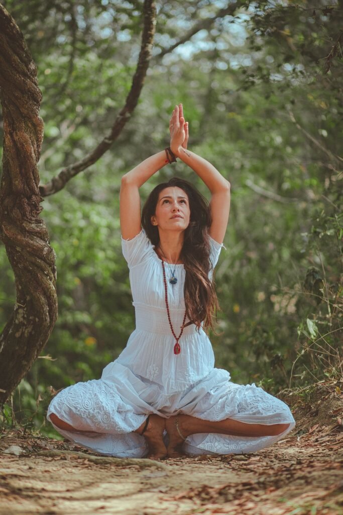 Mulher meditando sentada em postura de lótus na floresta, com as mãos elevadas em mudra acima da cabeça, olhos voltados para cima em estado contemplativo
