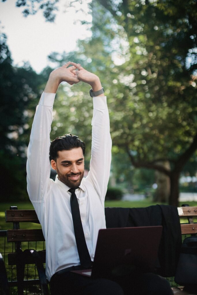 Homem de camisa social alongando os braços ao ar livre, ao lado de um notebook em um banco de parque