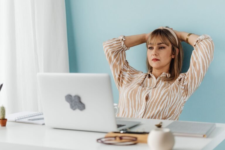 Mulher alongando o pescoço com as mãos atrás da cabeça em frente ao laptop, em pausa durante o trabalho no home office