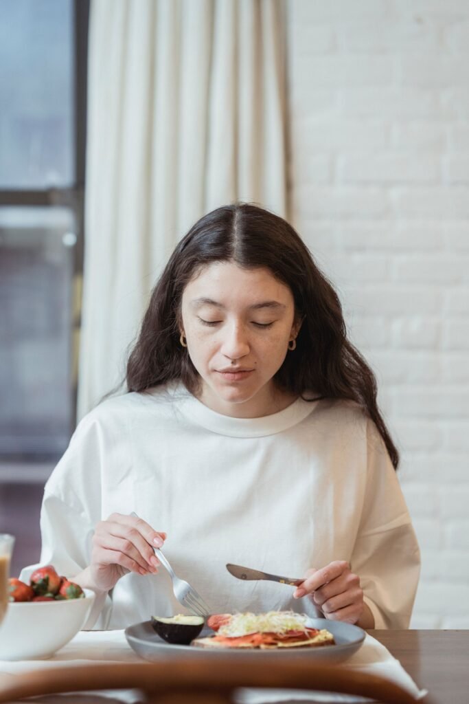 Mulher comendo com atenção plena, olhos fechados, saboreando uma refeição saudável com pão integral, abacate e morangos à mesa