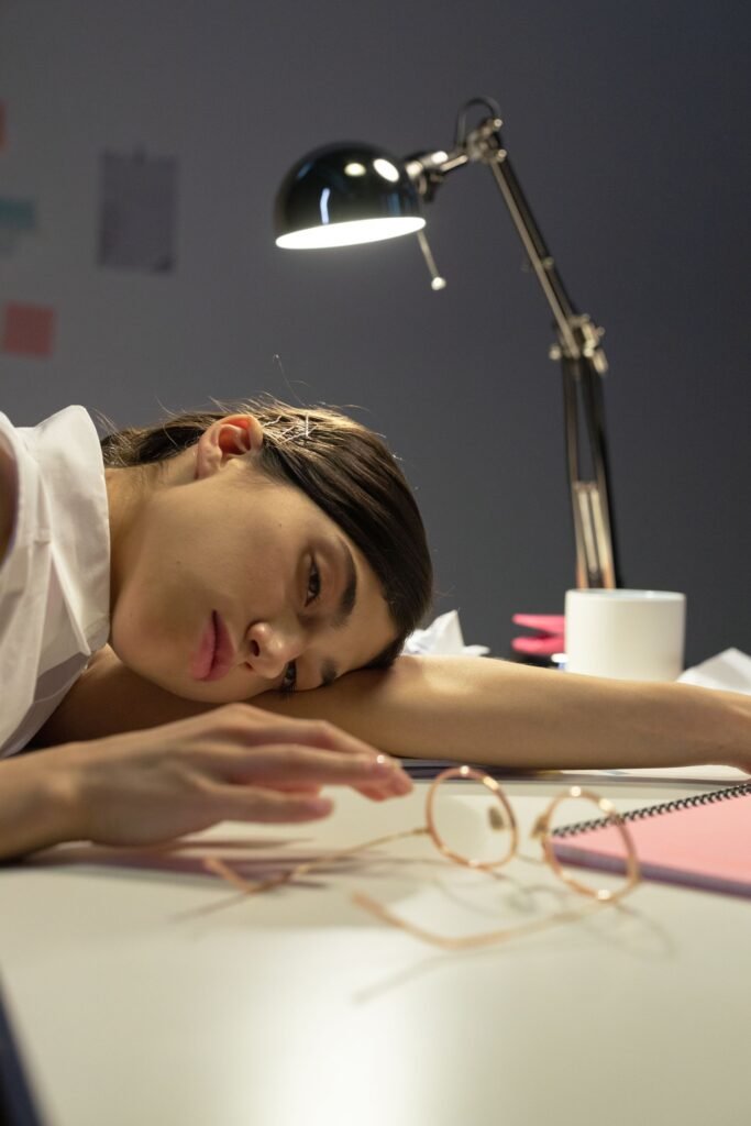 Mulher exausta com a cabeça apoiada na mesa de trabalho, cercada por óculos, caderno e luminária, representando o esgotamento do home office