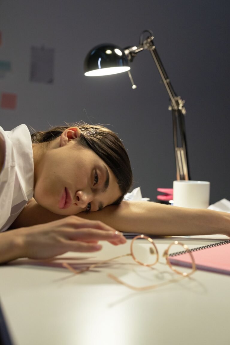 Mulher exausta com a cabeça apoiada na mesa de trabalho, cercada por óculos, caderno e luminária, representando o esgotamento do home office
