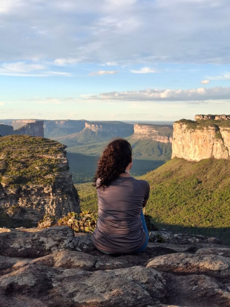 Mulher sentada de costas em formação rochosa, contemplando paisagem de montanhas e vale ao entardecer