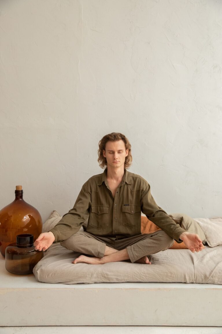 Homem sentado em posição de meditação sobre a cama, pernas cruzadas, mãos apoiadas nos joelhos, olhos fechados