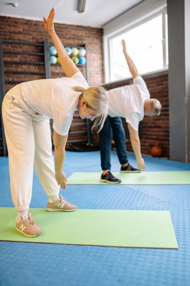 Casal de idosos praticando alongamento lateral de yoga em estúdio, sobre tapetes verdes